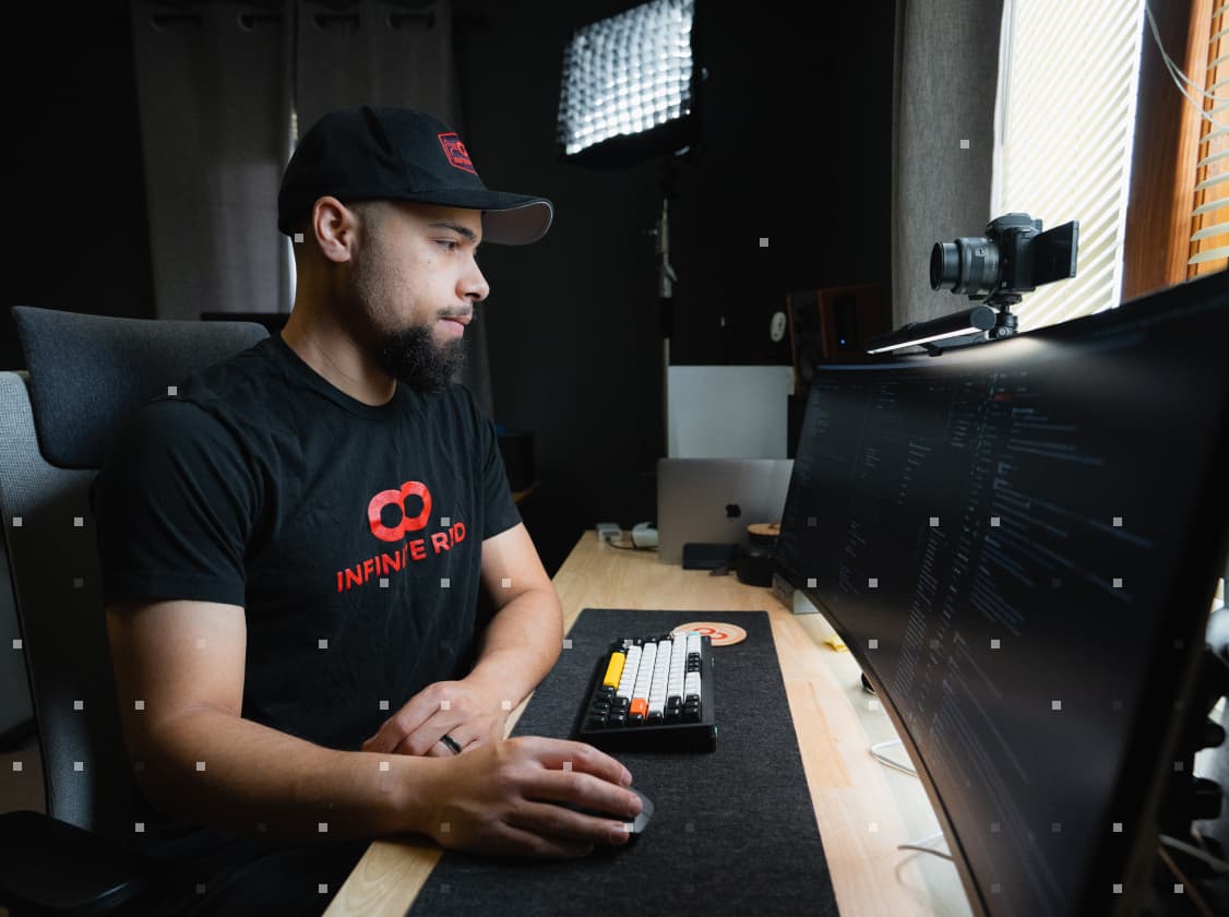 Infinite Red developer, Jon Major, focused on coding at his workspace. He is wearing a black Infinite Red T-shirt and a matching cap, seated in front of a computer with code visible on the screen, and video recording equipment set up nearby.